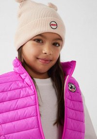 Roze doorgestikt vest met ritssluiting en logo-patch, gedragen over een wit shirt, gecombineerd met een lichtroze geribbelde muts met ooraccenten en logo.