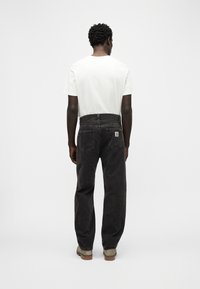 Άνδρας που στέκεται με την πλάτη στραμμένη, φορώντας λευκό μπλουζάκι, μαύρο τζιν με πίσω τσέπες και ανοιχτό καφέ παπούτσια σε λευκό φόντο.