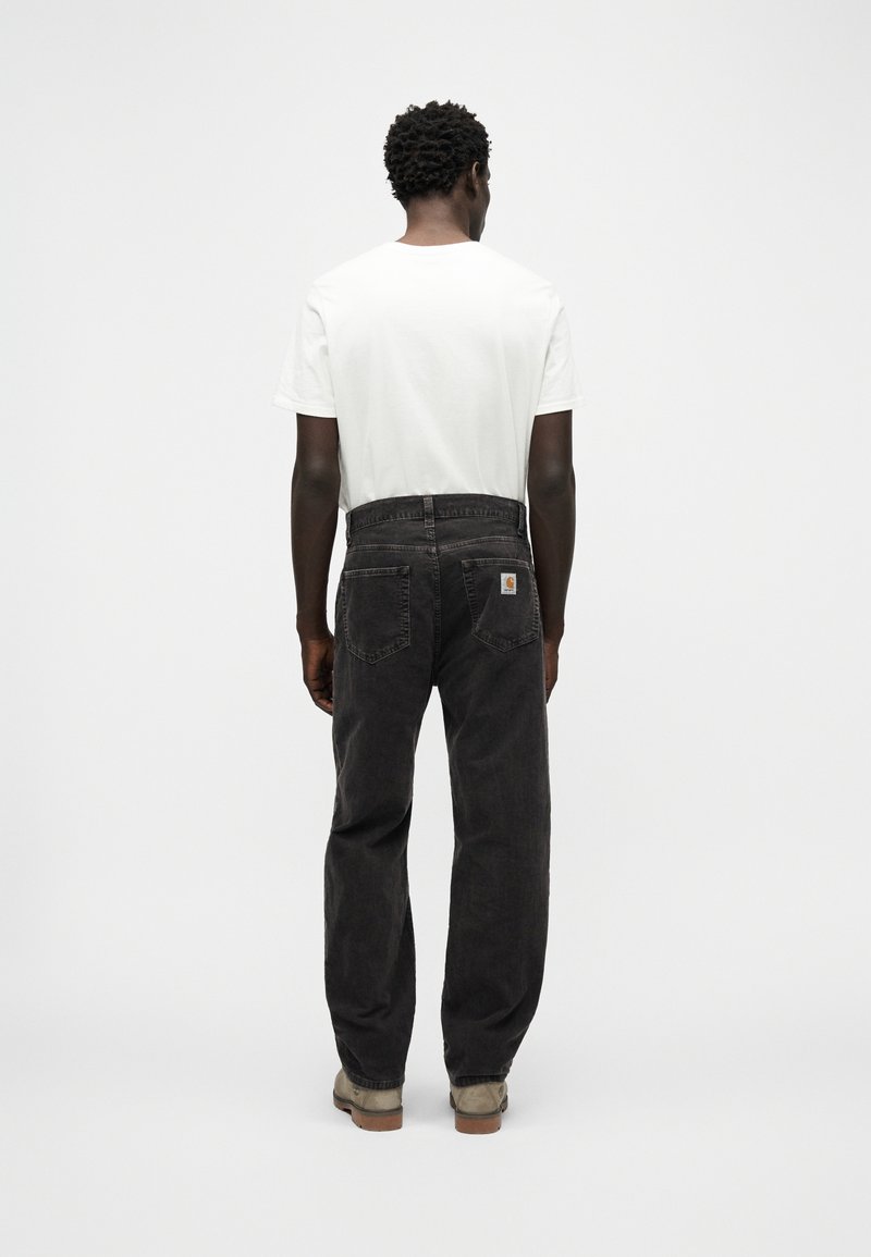 Homme debout de dos portant un t-shirt blanc, un jean noir avec des poches arrière, et des chaussures brun clair sur un fond blanc.