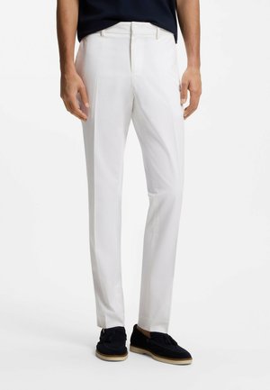 Homme portant un pantalon blanc slim et des mocassins noirs en daim avec pompons, debout devant un fond clair uni.