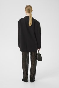 Blazer nero oversize, vestibilità ampia, pantaloni in rete nera texturizzati, dettagli lucidi, tiene in mano una piccola borsa nera, design minimalista.