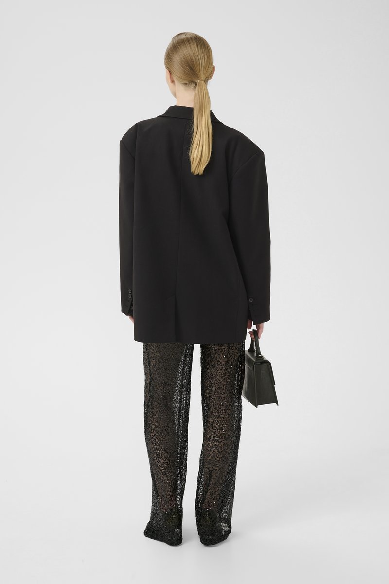 Overdimensioneret sort blazer, løst fit, teksturerede sorte mesh-bukser, skinnende detaljer, holder en lille sort håndtaske, minimalistisk design.