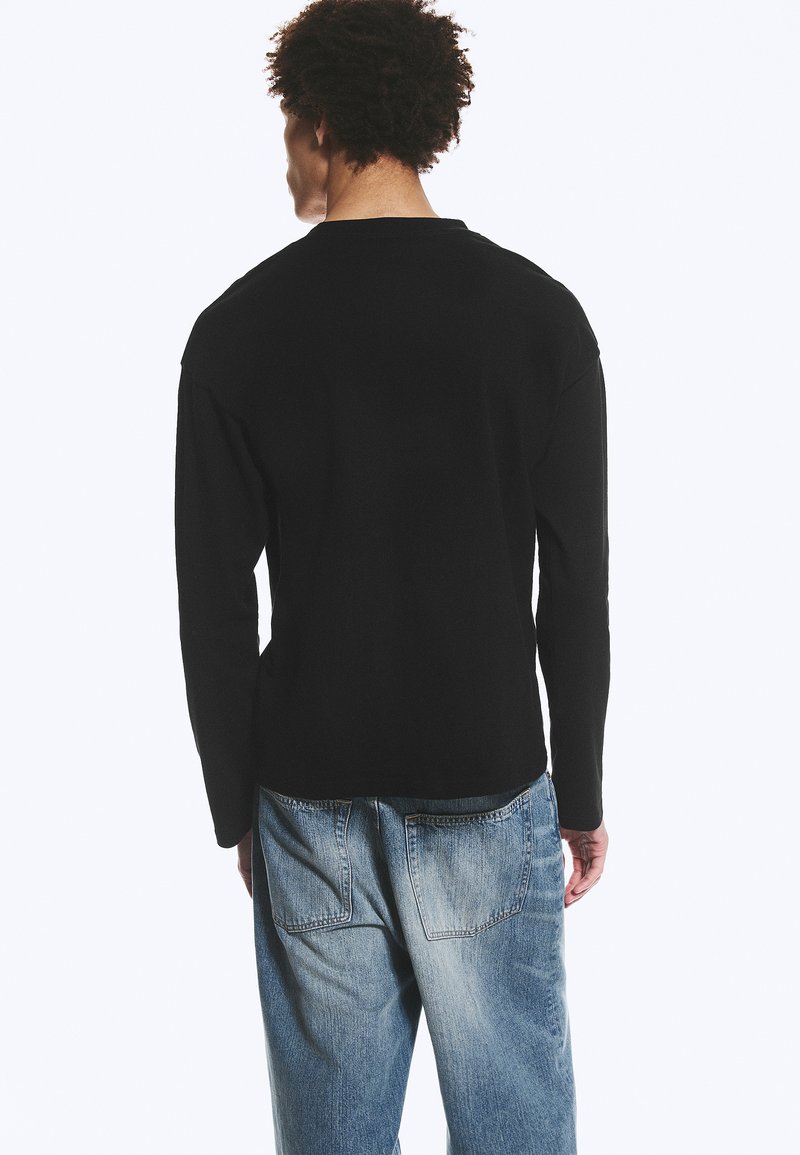 Uomo visto di spalle che indossa una maglietta nera a maniche lunghe semplice e jeans azzurri chiari, larghi, su sfondo bianco.