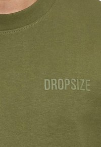 Olivegrünes T-Shirt mit Rundhalsausschnitt und einem strukturierten Druck des Wortes "DROPSIZE" in einer helleren Nuance. Glattes, aus Baumwolle gefertigtes Material.
