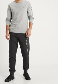 Graues Langarmshirt mit einem Logo auf dem Ärmel, kombiniert mit dunkelgrauen Jogginghosen mit weißem Text und schwarzen Sneakern. Einfaches Design.