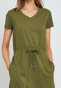 Robe courte olive en tissu doux, à manches courtes, avec un col en V, une taille froncée avec cordon de serrage et des poches latérales.