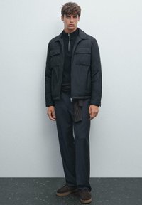 Black zip-up jacket with two chest pockets, paired with a ribbed black turtleneck and dark trousers. Brown footwear completes the look.