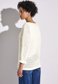 Weißer, locker gestrickter Pullover mit Streifenmuster, runder Ausschnitt und Drei-Viertel-Ärmeln, kombiniert mit blauen, weit geschnittenen Jeans.