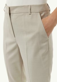 Beżowe spodnie o fakturze z gładkim wykończeniem, bocznymi kieszeniami i subtelnymi detalami szwów w okolicy talii. Materiał wydaje się lekki i przewiewny.