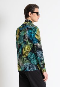 Camisa de manga corta en negro con un vibrante patrón de hojas en verde, azul y amarillo. Presenta un cuello clásico y un diseño de ajuste slim.