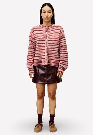 Vrouw in een roze en rode gestreepte knoopcardigan, donkerrode leren minirok, kastanjebruine sokken en beige schoenen, staande tegen een witte achtergrond.
