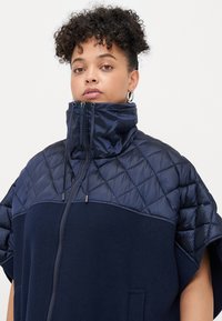 Veste à capuche marine avec une partie supérieure matelassée. Elle présente un col haut, une fermeture éclair et des poches latérales. Texture lisse et coupe décontractée.