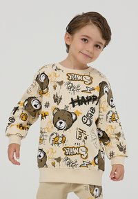 Sweatshirt beige con grafiche di orsi in stile cartoon, doodles astratti e testo. Tessuto morbido con polsini e orlo a coste. Maniche lunghe, vestibilità rilassata.