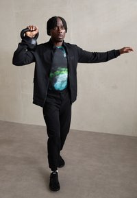 Schwarze Sportjacke, bunte Hemd mit abstraktem Muster, schwarze Jogginghosen, hält eine Kettlebell, gestylt mit schwarzen Sneakers in einem minimalistischen Setting.