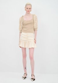 Top in maglina beige con maniche trasparenti a sbuffo, abbinato a una mini gonna di raso crema con volant. Le calzature presentano un design a stampa bovina in bianco e nero.