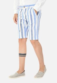 Pantaloni corti in lino a righe blu e bianche con vita regolabile tramite cordino, dotati di tasche laterali e dettaglio con bottone. Scarpe slip-on casual di colore grigio chiaro.