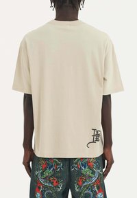 Camiseta de manga corta color beige con escote redondo, que presenta un diseño gráfico negro en la parte inferior de la espalda. El material parece suave y liso.