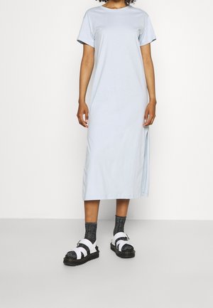 Personne portant une robe midi bleu clair à manches courtes, associée à des chaussettes noires et des sandales blanches à boucle sur un fond uni.