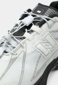 Scarpa sportiva bianca con sovrapposizione testurizzata, lacci integrati e marchio visibile. Presenta una suola nera e accenti grigi.