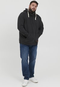 Svart hettegenser med beige snorer, kengurulomme, ribbede mansjetter, sammen med mørkeblå jeans og hvite sneakers.