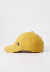 Gul bomull baseballkeps med en böjd skärm, försedd med en broderad svart "CK"-logga på framsidan och ventilationshål på sidorna.