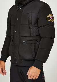 Sort dunjakke med tekstureret finish, trykknapper, to forlommer og et mærke på venstre ærme. Ribstrikkede manchetter fuldender designet.