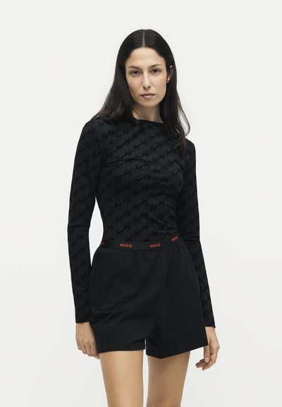 Femme aux longs cheveux foncés portant un haut à manches longues noir à motifs et un short noir avec une ceinture rouge avec le logo "HUGO".