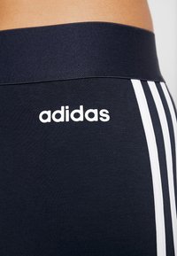 Navy leggings with a wide, elastic waistband; features white Adidas logo and signature three stripes along the sides. Smooth fabric texture.