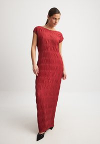 Red, form-fitting, maxi dress with vertical ruching, cap sleeves, and a smooth texture. Accented with pointed black shoes.