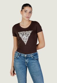 Camiseta de algodón marrón con mangas cortas, con un logo triangular plateado y negro con el diseño "GUESS" en el frente. Combinada con vaqueros azules.