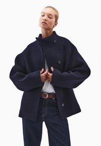 Ciemnoniebieska marynarka z wełny z wysokim kołnierzem, du�żymi guzikami i szerokimi rękawami. Połączona z białą bluzką i ciemnymi dżinsami.