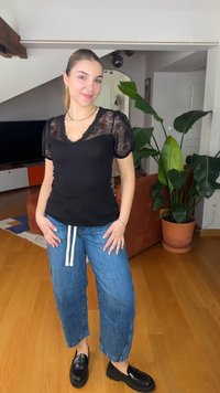 Jonge vrouw staat glimlachend in een casual zwart kanten top, blauwe spijkerbroek en zwarte loafers in een woonkamer met een houten vloer en een grote potplant.