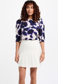 Witte geplooide rok met knoopaccenten en een losse, bedrukt top met grote paarse en blauwe bloemdesigns op een lichte achtergrond.