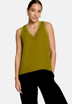 Femme portant un haut sans manches col en V vert olive et un pantalon noir à jambes larges sur un fond clair uni.