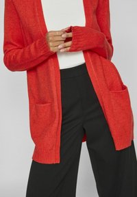 Röd stickad kofta med ribbade manschetter och två framfickor, i kombination med svarta vida byxor och en vit topp. Mjuk textur.
