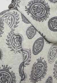 Šedá mikina s kapucí se vzorem opakujících se tmavě modrých emblémů. Vyrobena z měkké látky s texturovaným povrchem, obsahuje kapuci se stahovací šňůrkou.