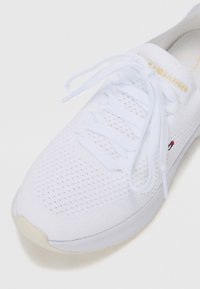White athletic shoe with a breathable mesh upper, flat laces, and a rubber sole. Features branded text in gold and small logo accents.