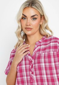 Camicia a quadri rosa con maniche corte, dotata di colletto a button-down e tasca frontale. Realizzata in tessuto leggero con una vestibilità rilassata.