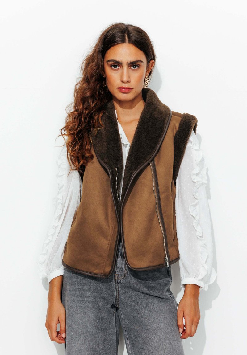 Gilet marron sans boutons, avec un col en peluche et une fermeture éclair à l'avant. Porté par-dessus une chemise blanche à manches longues avec volants, associé à un jean gris.