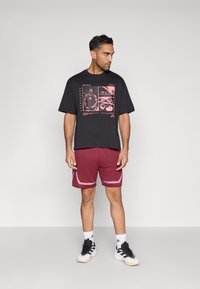 Must graafiline T-särk roosade mustritega, kombineeritud burgundia punaste lühikeste pükstega, millel on valged äärised. Modell kannab musta-valgeid spordijalatseid valgete sokkidega.