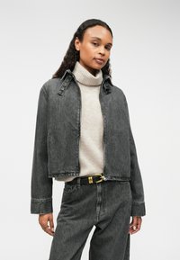 Szara dżinsowa kurtka o skróconym fasonie, z kołnierzykiem na zatrzaski i widocznymi szwami. Noszona na jasnobeżowym swetrze z golfem.