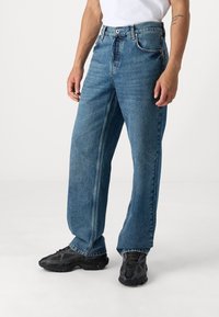 Muž v volných modrých džínách a černých sportovních botách, stojící proti jednobarevnému světlému pozadí.