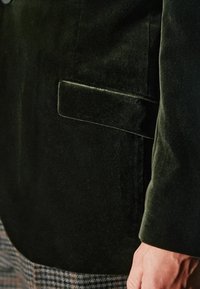 Ciemnozielona aksamitna marynarka z wyraźnym kołnierzem, pojedynczą kieszenią na piersi oraz teksturowanym materiałem, w połączeniu z wzorzystymi spodniami.