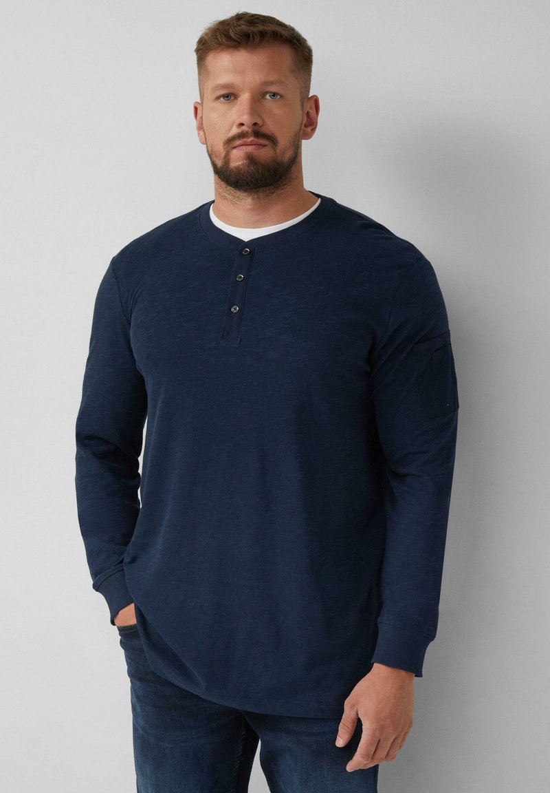 Marineblaue Langarm-Henley-T-Shirt mit einem weißen Kragen, einer Knopfleiste mit drei Knöpfen und einer Tasche am linken Ärmel. Glatte Textur, lockere Passform.
