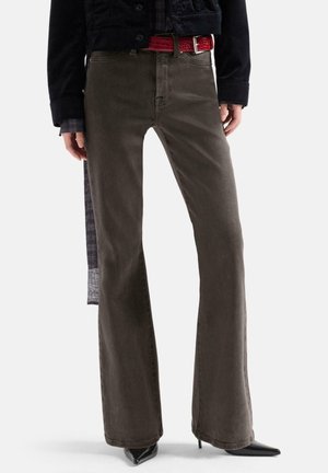 Persona che indossa pantaloni marrone scuro a gamba larga, scarpe nere con tacco alto appuntite, una cintura rossa e una giacca nera su sfondo uniforme.
