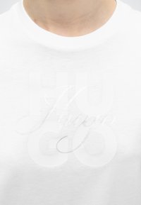 Κοντινή λήψη του λαιμού ενός λευκού T-shirt με διακριτικό ανάγλυφο κείμενο που γράφει "Hugo" και είναι τοποθετημένο πάνω από μεγάλα πεζογράμματα.