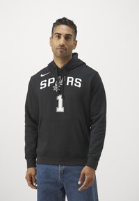 Fekete kapucnis pulcsi, fehér színű "SPURS" felirattal és "1" számmal. Kanguru zsebe van és kengyeles kapucnija. Kék farmernadrággal viselve.