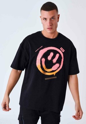Camiseta negra de gran tamaño hecha de algodón, que presenta un gran gráfico de una carita sonriente en colores rosa y naranja, con texto impreso en la parte frontal.
