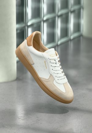 Biały i beżowy sneaker z jasnobrązową podeszwą i piętą, białymi sznurowadłami, logo "GANT" na boku i języku, unoszący się nad polerowaną betonową podłogą.