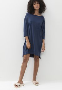 Marineblauw, losse jurk met driekwarts mouwen, een zak op de linkerborst en zijsplitten; gedragen met zachte, beige pantoffels.